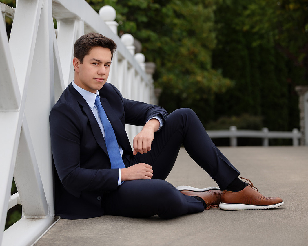 Senior guy in a navy suit seated on a bridge during senior pictures in Hamilton County, Indiana